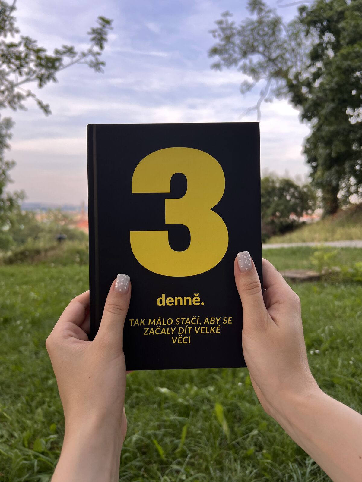 Deník 3 denně držený v rukou s výhledem na Prahu